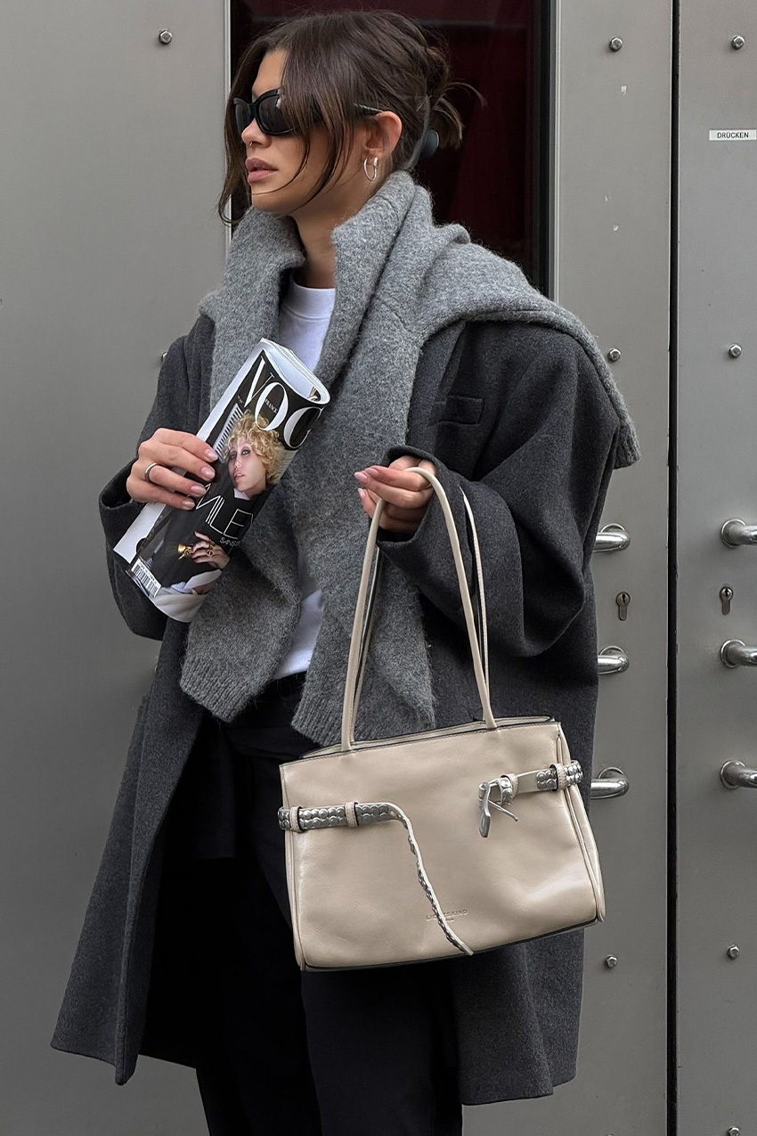 Berlin fashion trends are shown in a close street style photo of a woman standing outside in front of a silver door wearing black sporty sunglasses, hoop earrings, a gray cardigan draped over the shoulders styled with a gray boxy blazer, white t-shirt, black trousers, and a tan handbag adorned with a belt detail