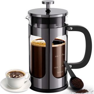 A black and glass french press with a cup of espresso coffee on a saucer and a black plastic scoop with ground coffee