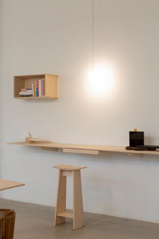 Interior view of the Formafantasma Milan studio featuring grey floors, white walls, a pendant light, a high wooden stool and a wall-mounted desk with a model and other items on it