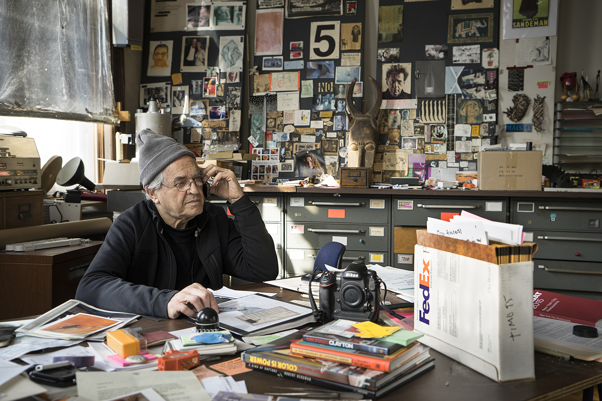 Jay Maisel wearing a grey beanie speaks on a cellphone while sitting at a cluttered desk filled with books, papers, and a Nikon camera, set against a wall covered in photos and art collages.