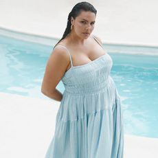 model wears blue shirred sun dress and white sandals with a pool in the background