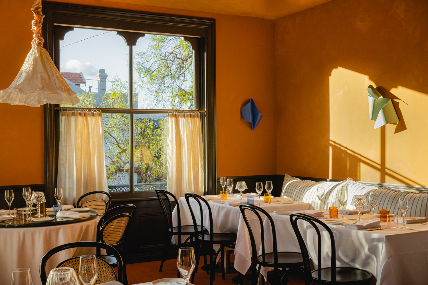 A ochre-washed restaurant bathing in sunshine boasts classic bistro tables mixed and matched with a quirky papier-mache-like chandelier in white, a sculptural sconce made of folded iron sheet, stripy cushions and transparent, blue, and yellow glassware.