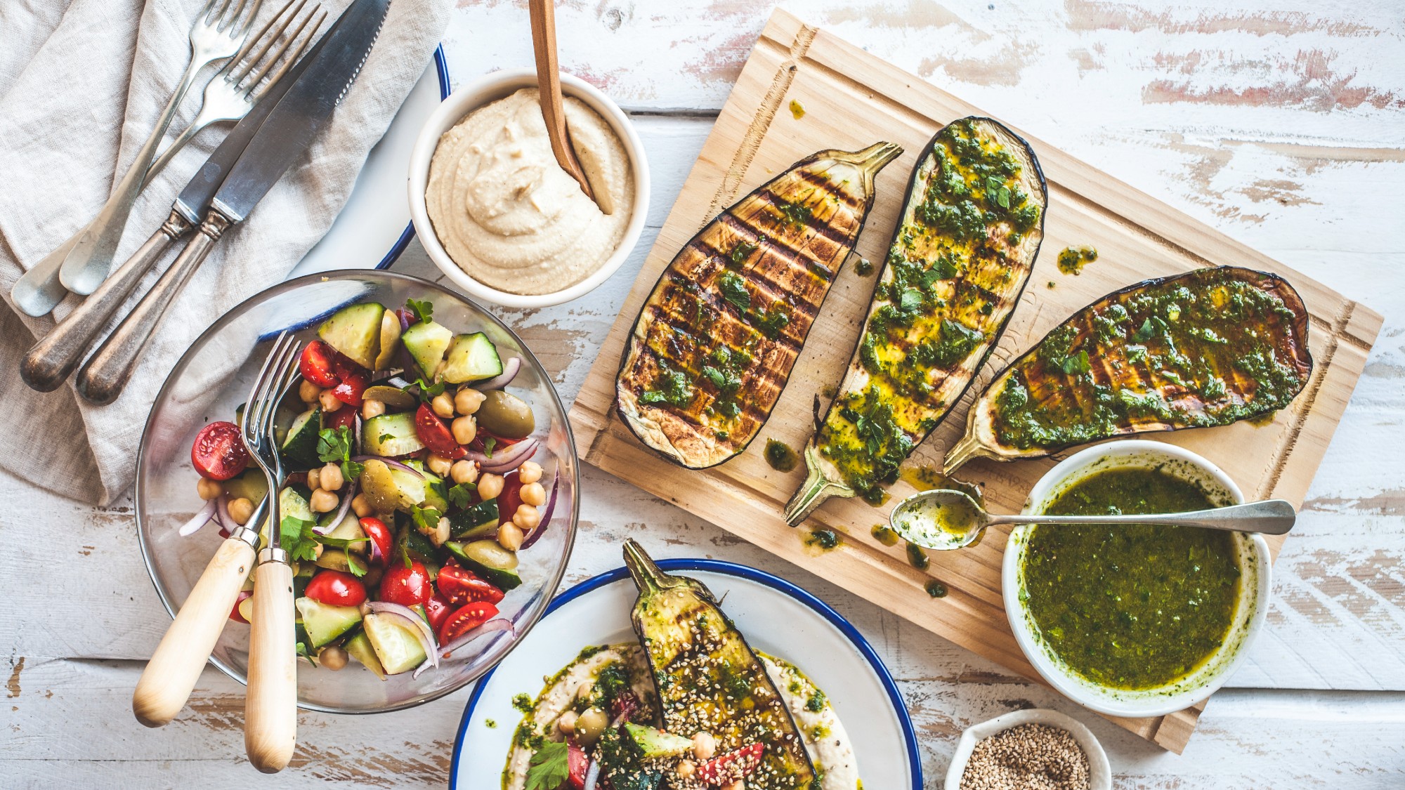 overhead shot of grilled eggplant on a cutting board along with a green sauce, hummus and a chickpea-cucumber salad