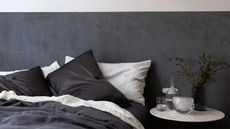 bed made with charcoal and pinstripe bedding with silk pillowcases and linen sheets, a marble side table with a carafe, glass, vase or flowers, and a dark velvet headboard behind the bed