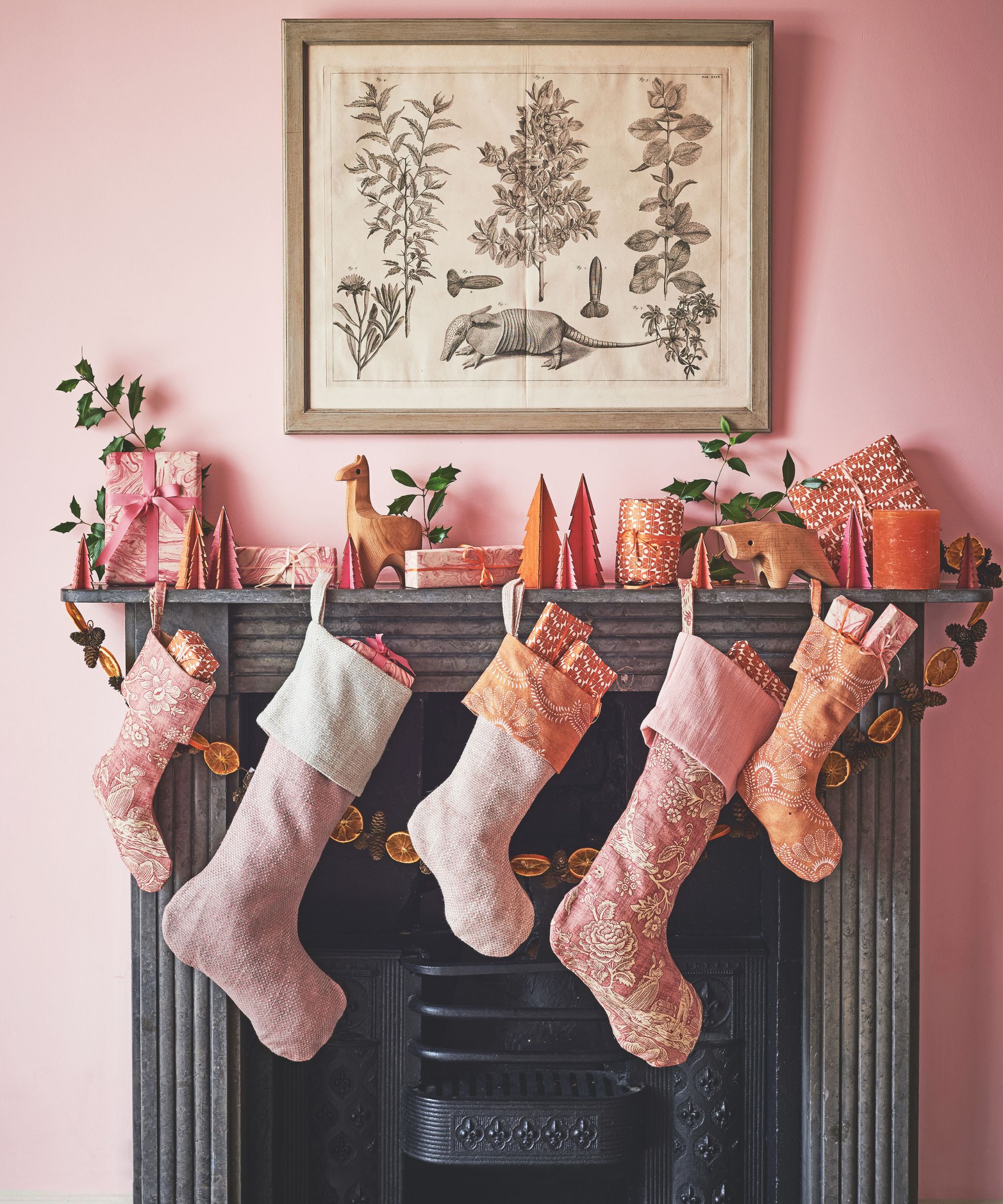 A festive mantelpiece in a pink room decorated with pink stockings, dried oranges, colorful trees, presents, and a botanical artwork above.