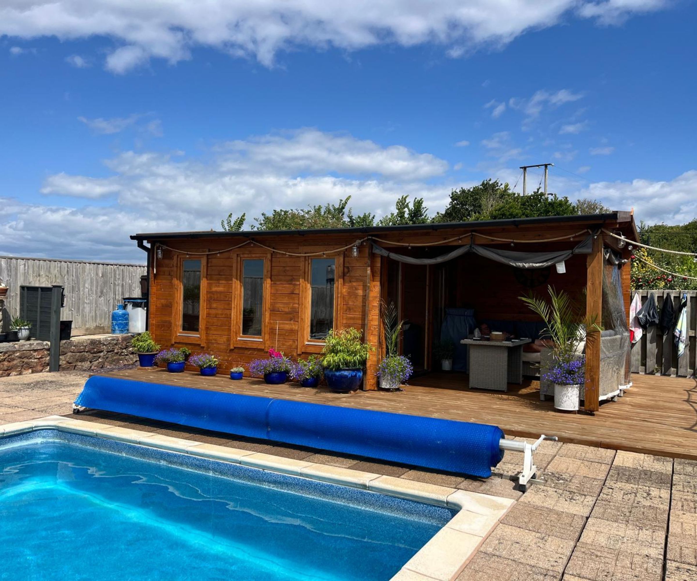 A timber log cabin pool house with an open-sided canopy, wicker garden furniture and blue potted flowers on a decked terrace, with a swimming pool and rolled solar cover in the foreground.