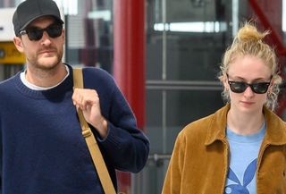 Sophie Turner with Peregrine Pearson at Heathrow Airport wearing a tan jacket, blue Playboy shirt, black leggings, and Ugg mules and carrying a Louis Vuitton bag.
