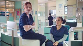 A younger Nicky look up at Jac as she sits on a desk in Holby City.
