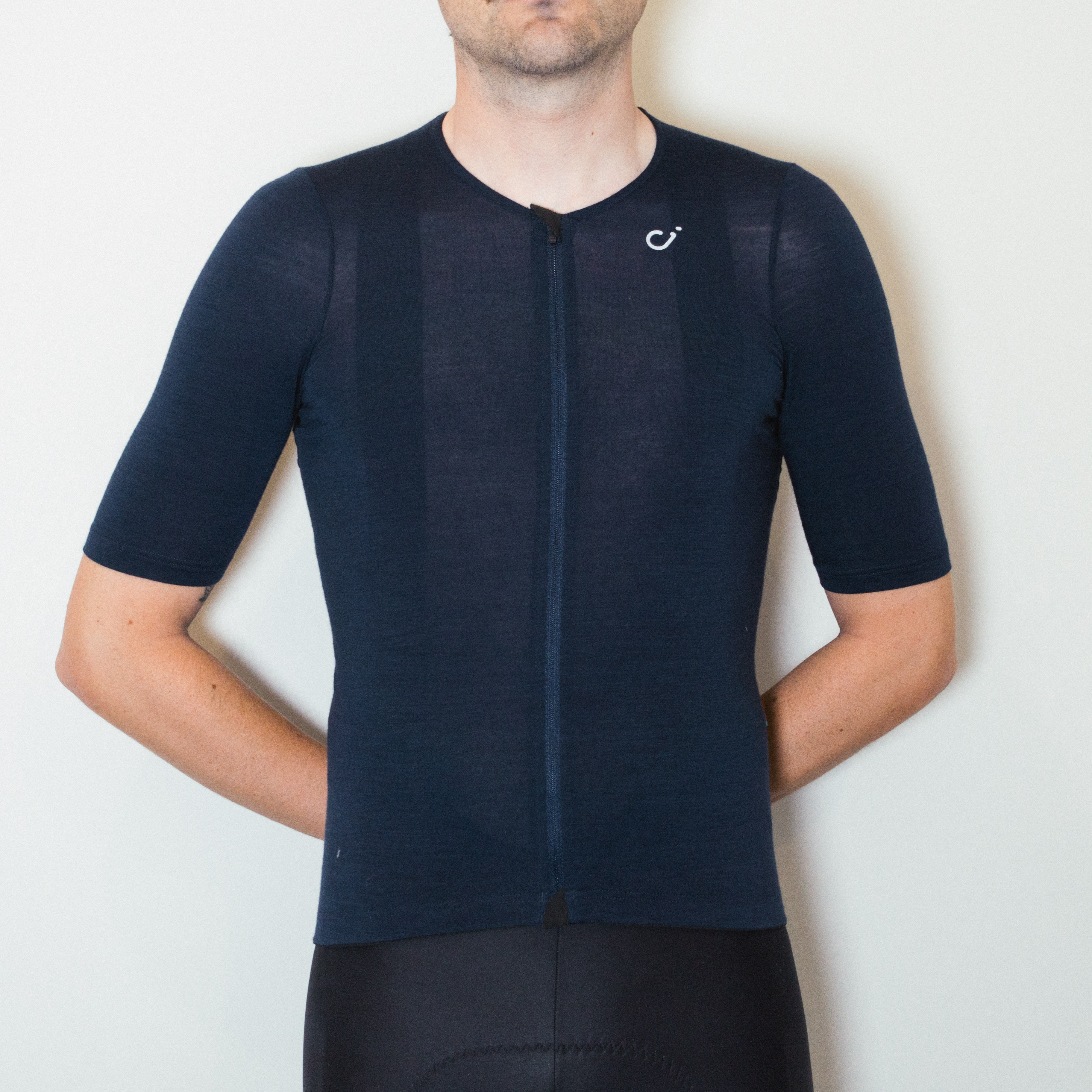 A white man stands against a white wall wearing black cycling shorts and a tight navy blue wool jersey