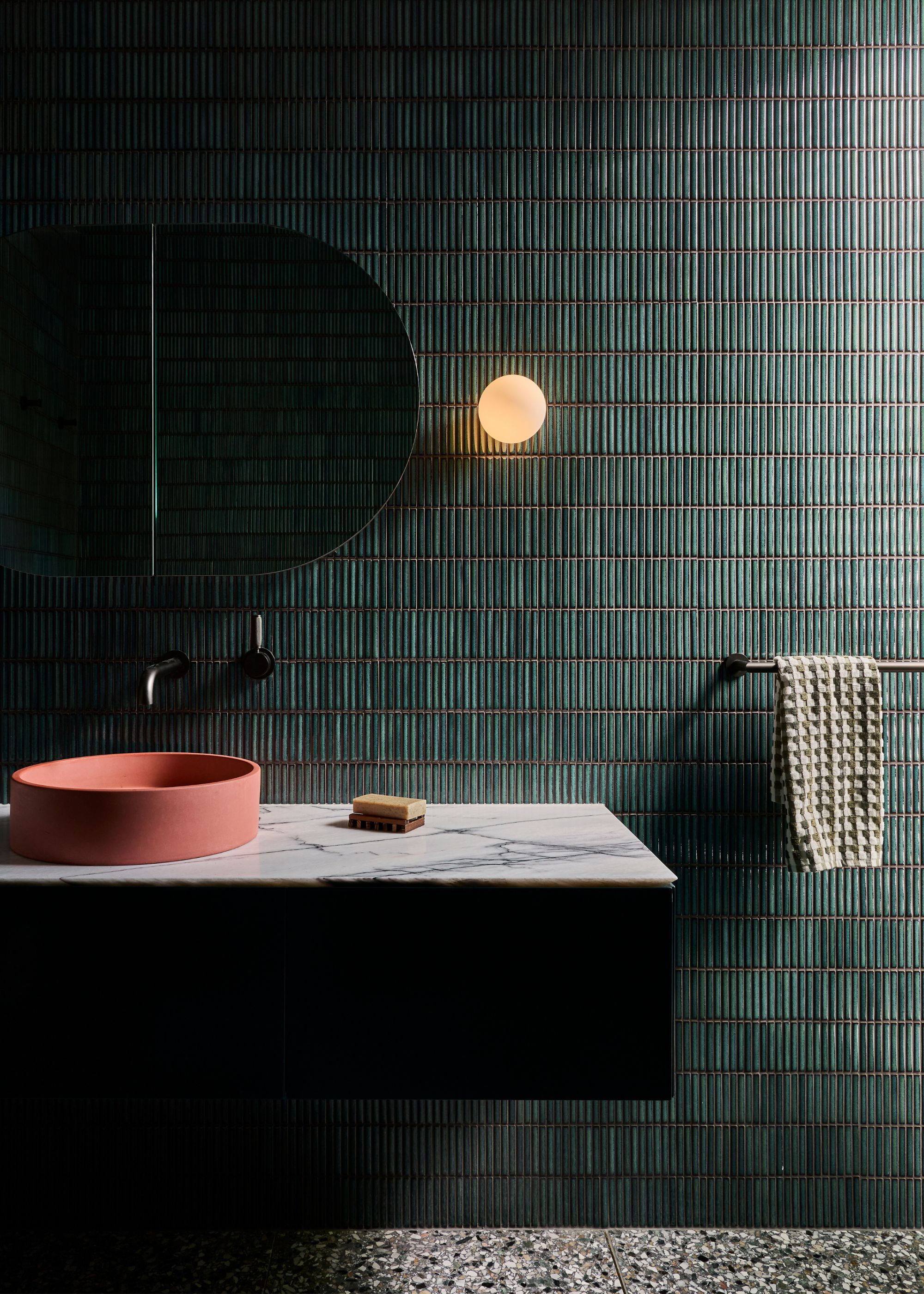 A green tiled bathroom, a single wall sconce, a terrazzo floor, a floating basin, and a coral sink