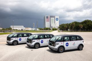 three electric minivans with nasa logos sit in a parking lot in front of a large building