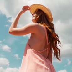 A woman with long, red hair wearing a pink strappy sun dress and a straw hat looking up to the sun in the sky