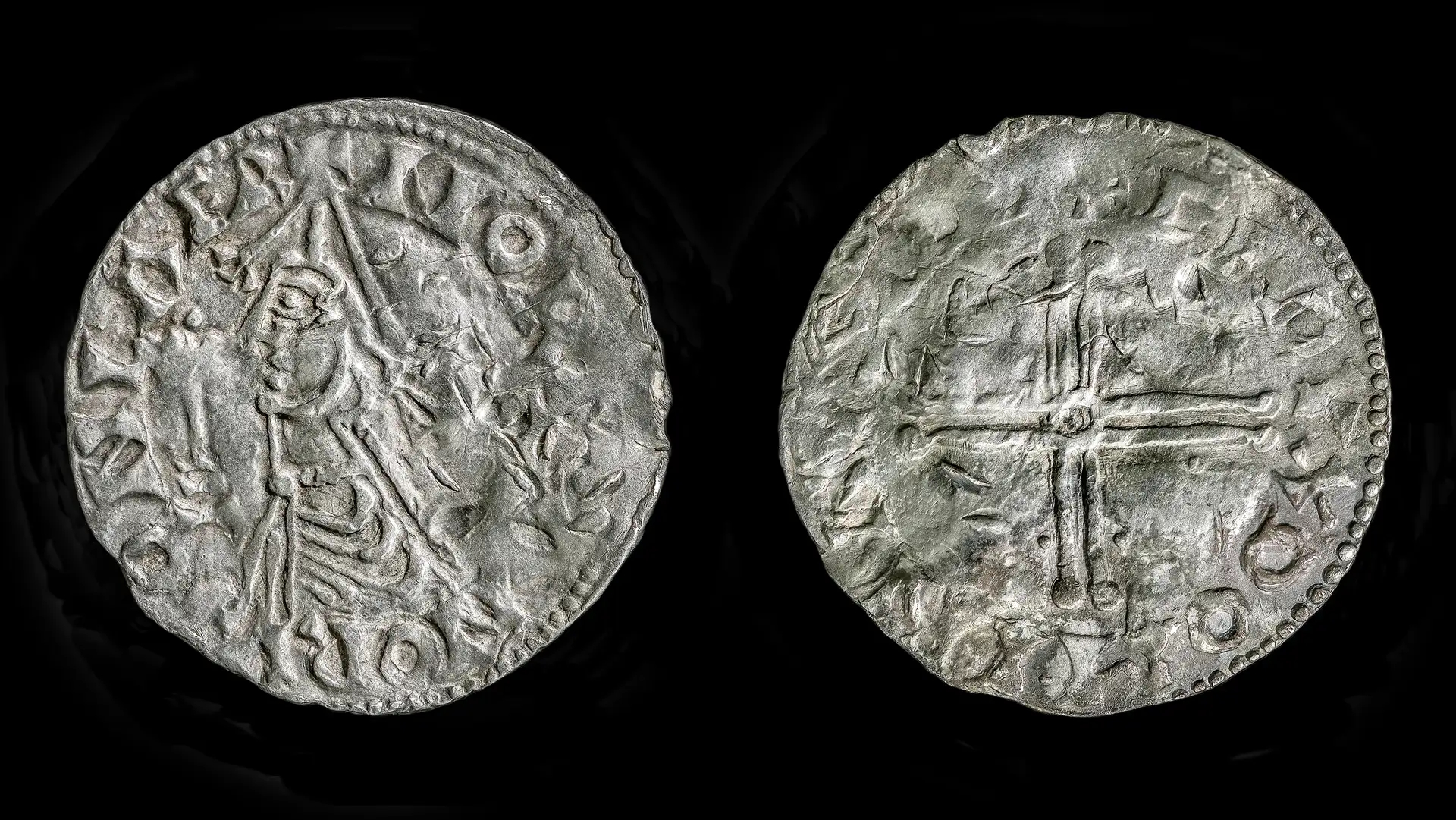 A close up of the back and front of a silver coin; one side shows a man's profile and the other shows a cross.