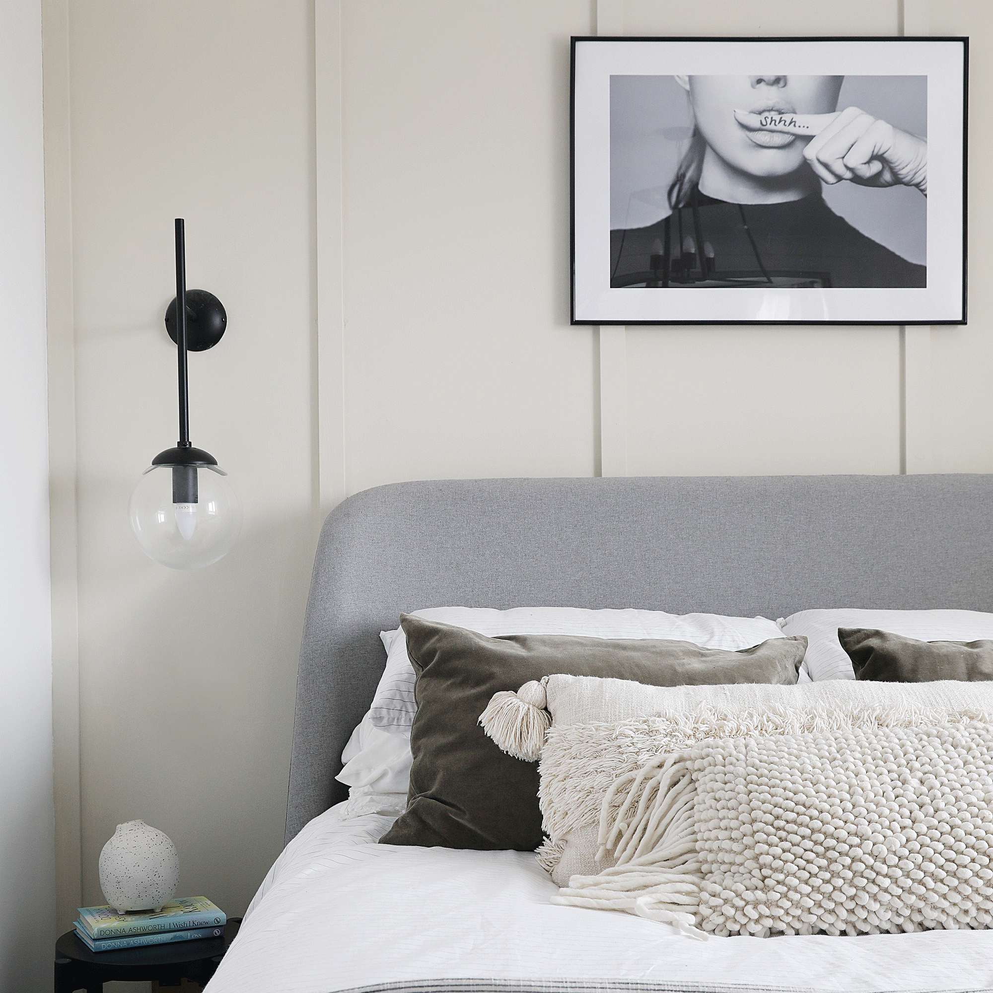 a neutral bedroom with beige panelled walls a grey upholstered headboard black globe wall lights black and white photo art