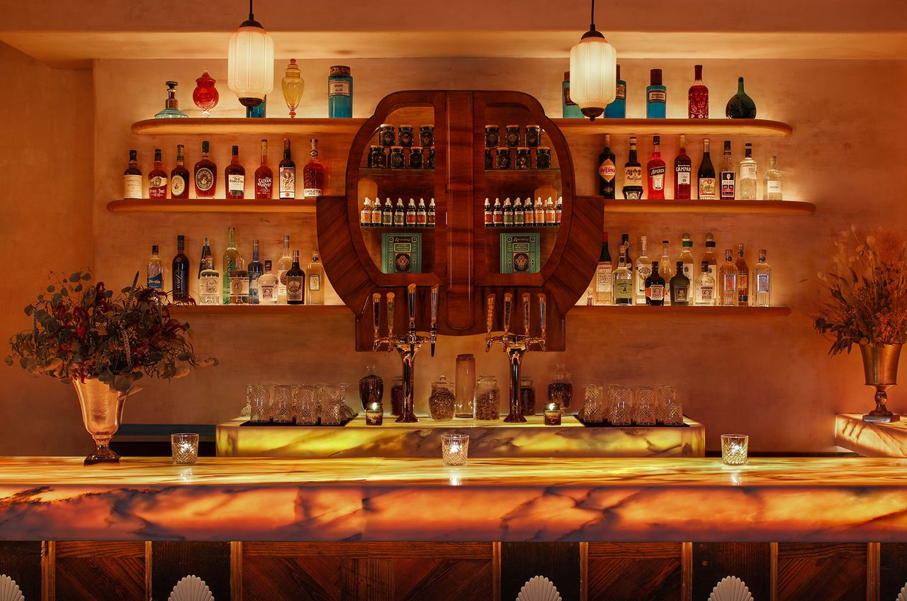 A Gatsbyan cocktail bar features marine green details that stand out against sleek woodwork, glass lighting accents, and the colorful selection of liqueurs stored behind the counter.