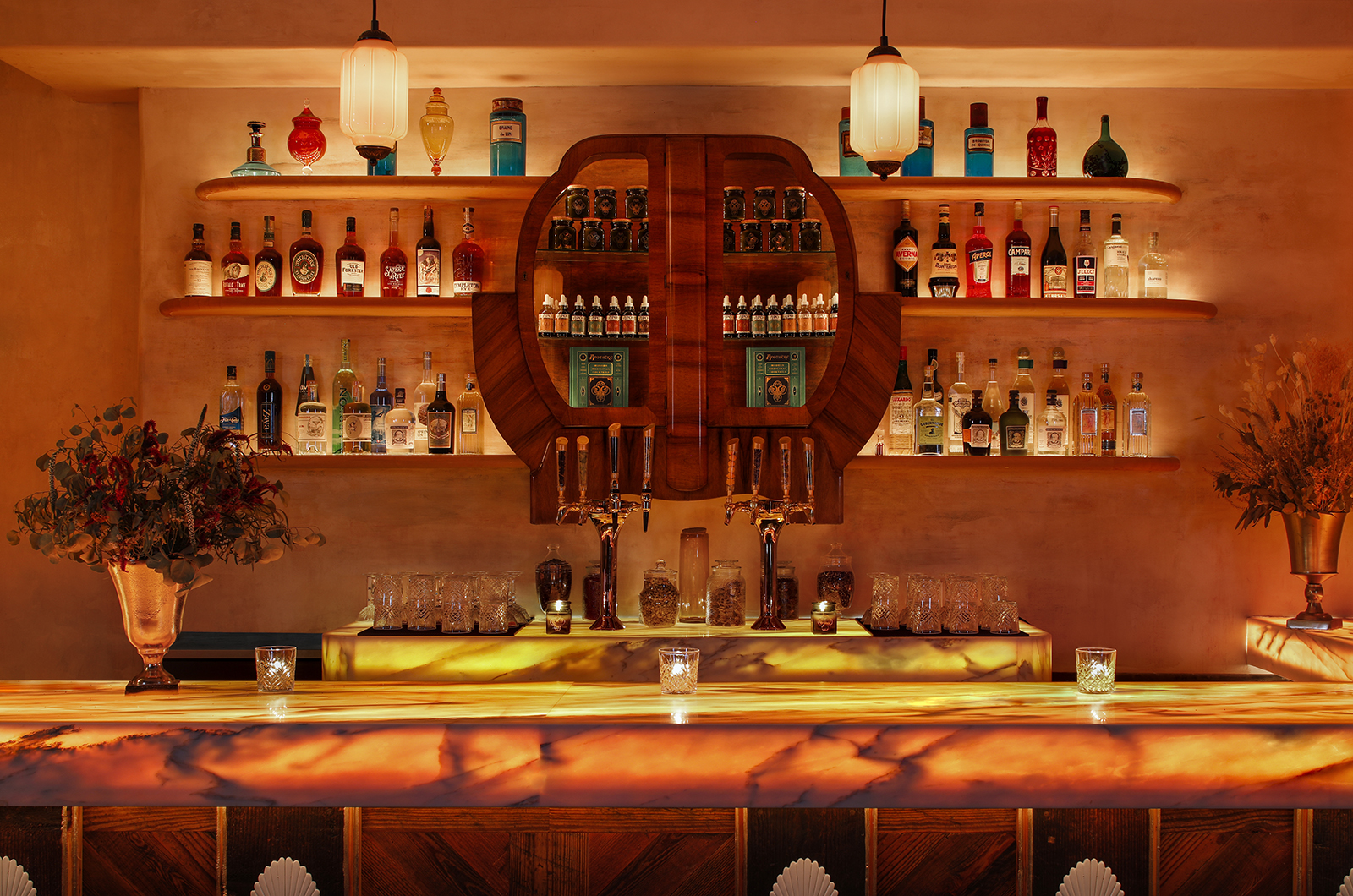 A Gatsbyan cocktail bar features marine green details that stand out against sleek woodwork, glass lighting accents, and the colorful selection of liqueurs stored behind the counter.