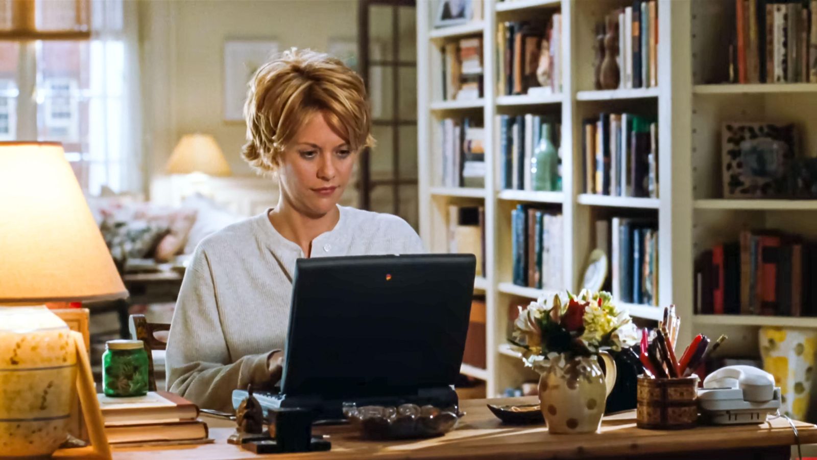 Meg Ryan in You&#039;ve Got Mail (1998) movie sat at a desk with a laptop with bookshelves behind her and a desk lamp