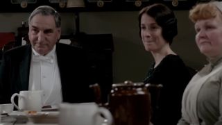 Jim Carter and co-stars sit at the downstairs table in conversation in Downton Abbey.