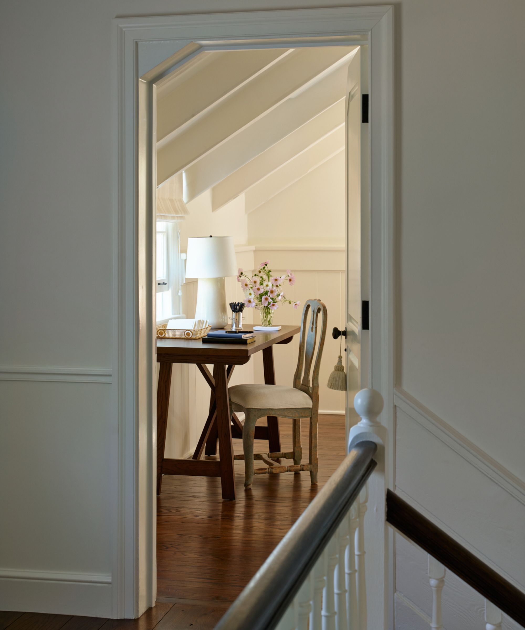 a peak through a doorway into a classic nancy meyers style bedroom with an antique wooden desk and chair