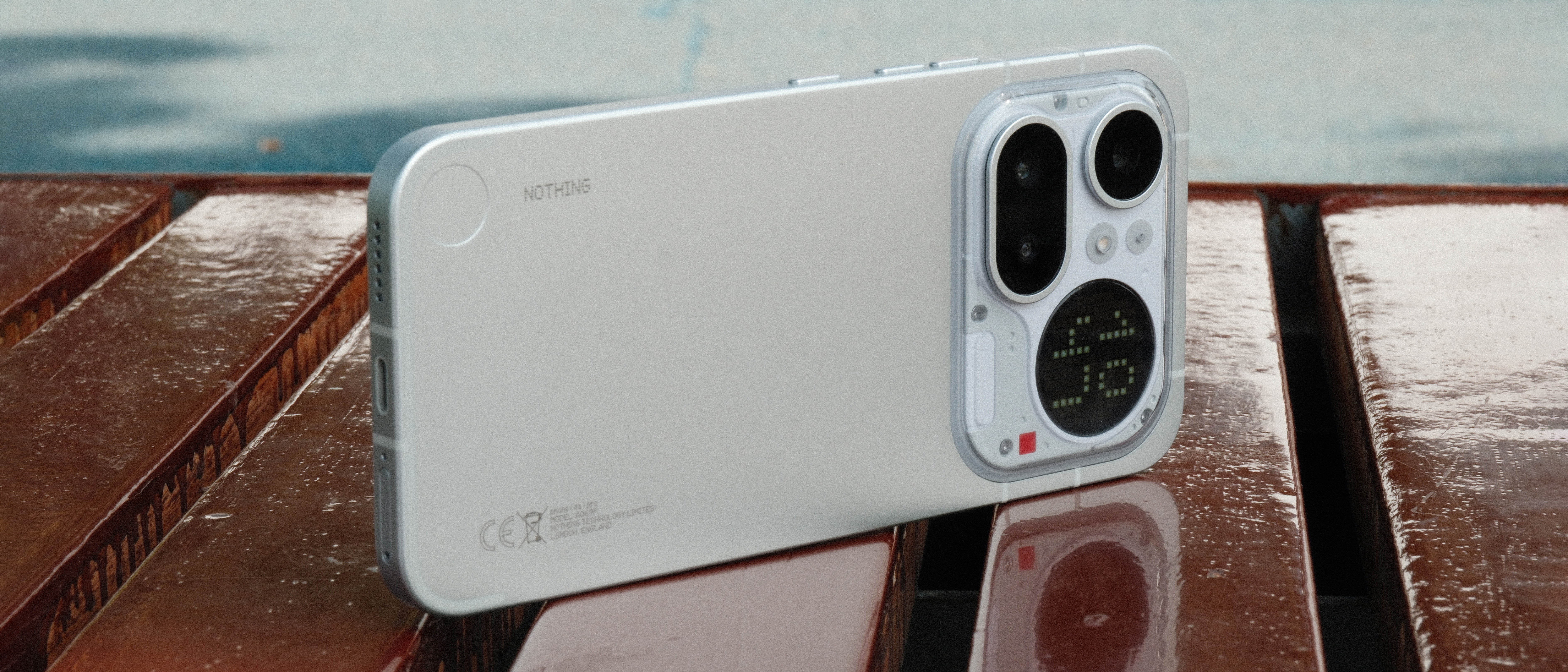 A light gray smartphone resting horizontally on a wet, reflective wooden bench. The back of the phone is visible, showcasing the camera island, the circular dot-matrix display, and a small red decorative square.