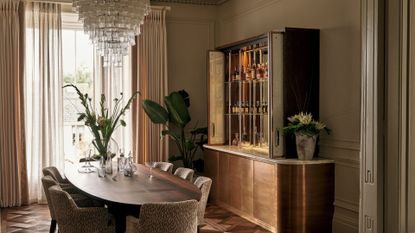 A beautiful dining room with a layered glass chandelier, a wood table, a patterned ding chair set, a sleek bar with a brushed brass base, a potted bird of paradise plant, and vases of flowers