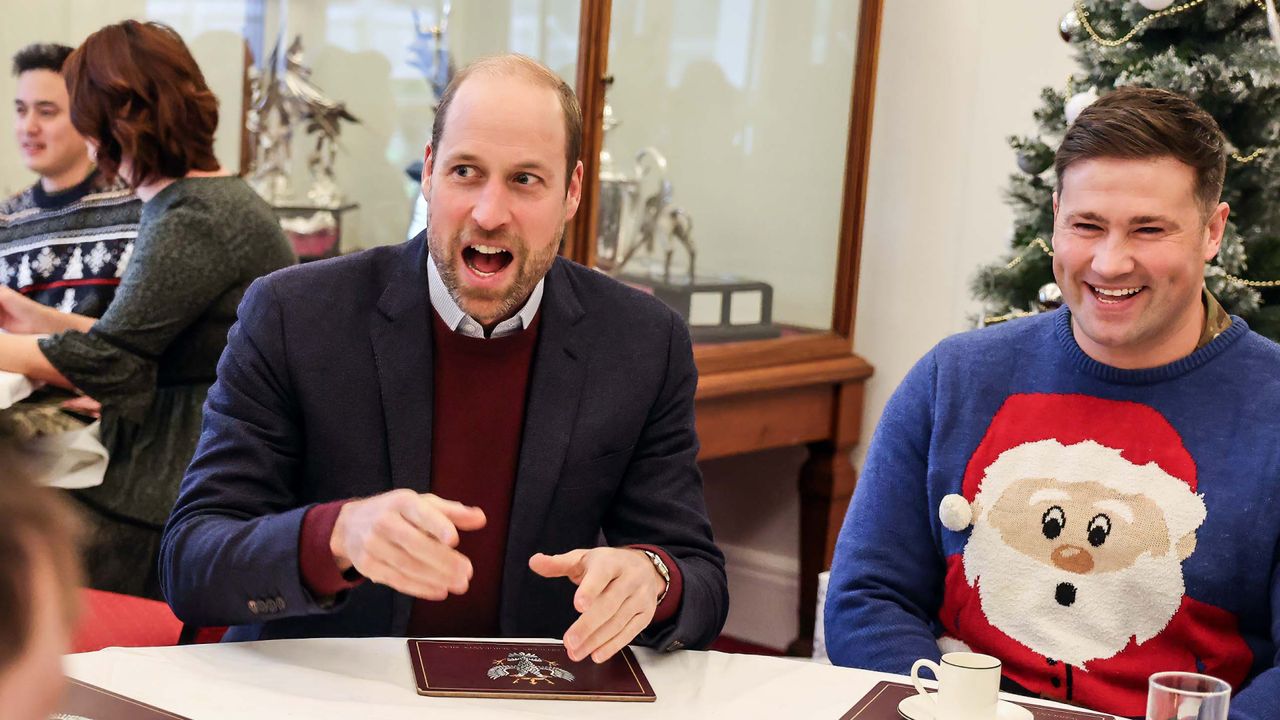 Prince William laughs while wearing a burgundy sweater and a suit jacket and sitting next to a man wearing a blue sweater with a big Santa face on the front