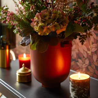 A floral arrangement with lit candles against a patterned wallpaper
