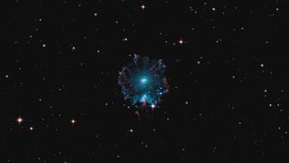 The Cat's Eye Nebula can be seen hanging against a backdrop of stars. The nebula has a wispy blue outer shell and a bright inner core.