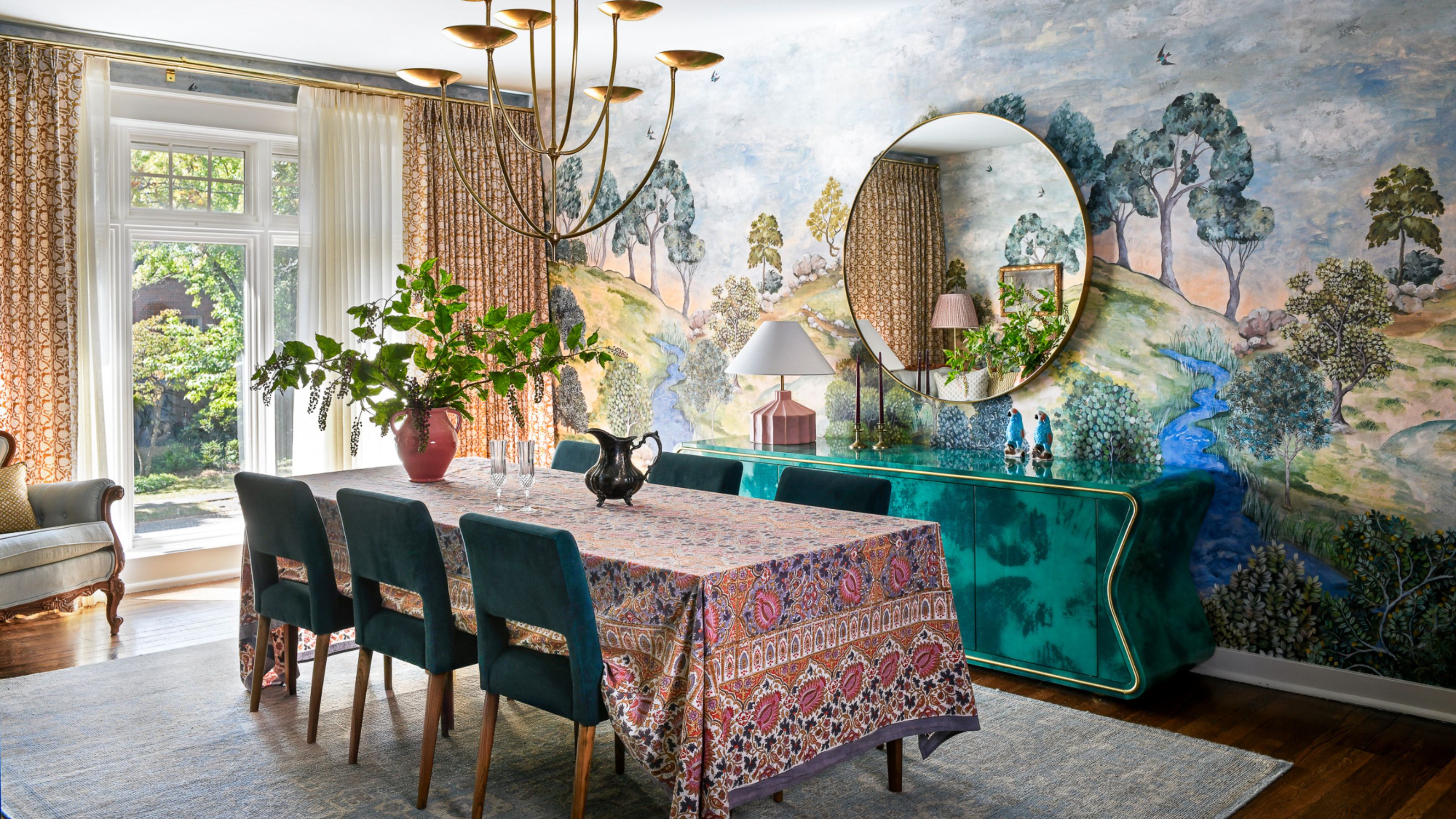 A colorful dining room with a forest scene wallcovering, a teal sideboard, and a dining table with teal chairs and a pink tablecloth.