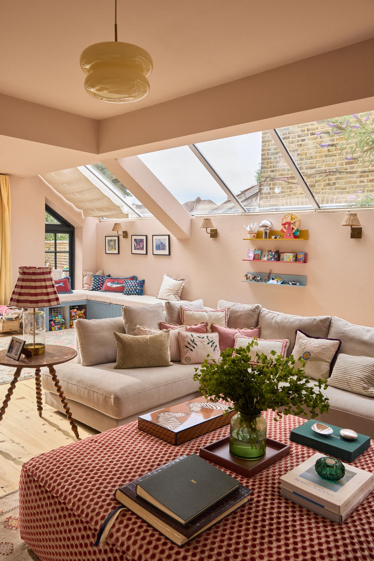 Image of a pink, traditional British-style living room with a large red patterned, upholstery ottoman, a sectional sofa with lots of pillows, and colorful decor.
