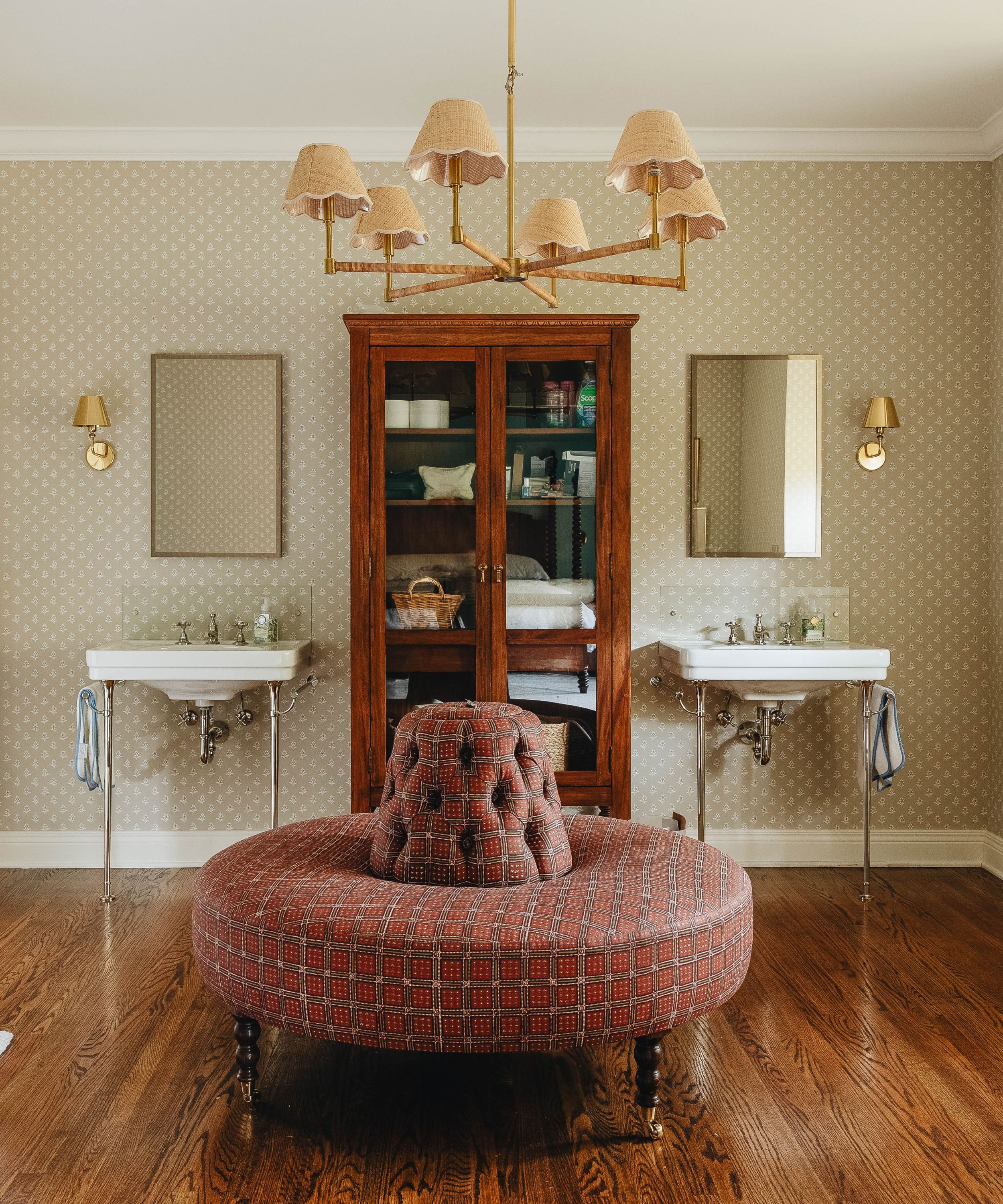 Traditional bathroom featuring patterned wallpaper, double pedestal sinks, a central wooden cabinet, and a large tufted red ottoman on hardwood floors.