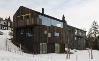 Alternative exterior view of the Oslo family house by STA - the house features a dark wood exterior, windows and doors with yellow frames and it is surrounded by other houses, trees and snow