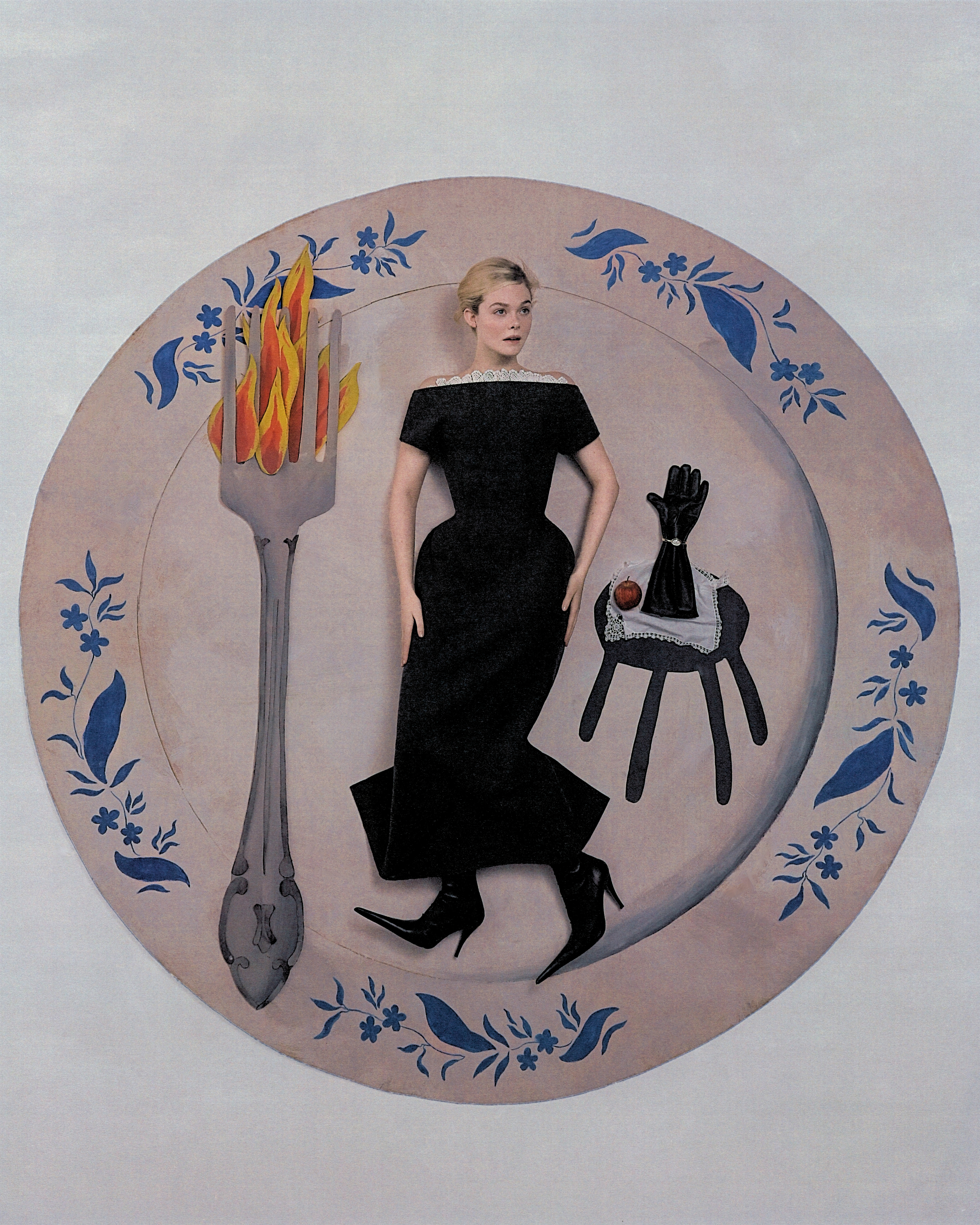 Elle Fanning shot by Szilveszter Mak&amp;oacute;. Fanning is lying flat on a life-size cardboard cutout of a dinner plate. She is wearing a black off-the-shoulder dress with black pointed-toe boots. On her left is a large cardboard cutout fork with flames, and on her right is a cardboard cutout side table with black glove and apple placed on top.