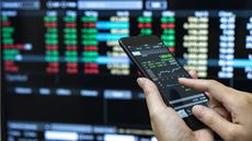 A woman, only her hands showing, holds a smartphone that shows stock trading on it with a backdrop of another display of stock trading.