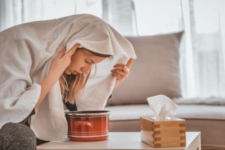 A woman holding her face over a bowl of hot water next to a box of tissues