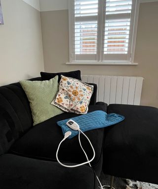 A blue heating pad on a black velvet sofa with assorted cream black and green cushions and shuttered window behind