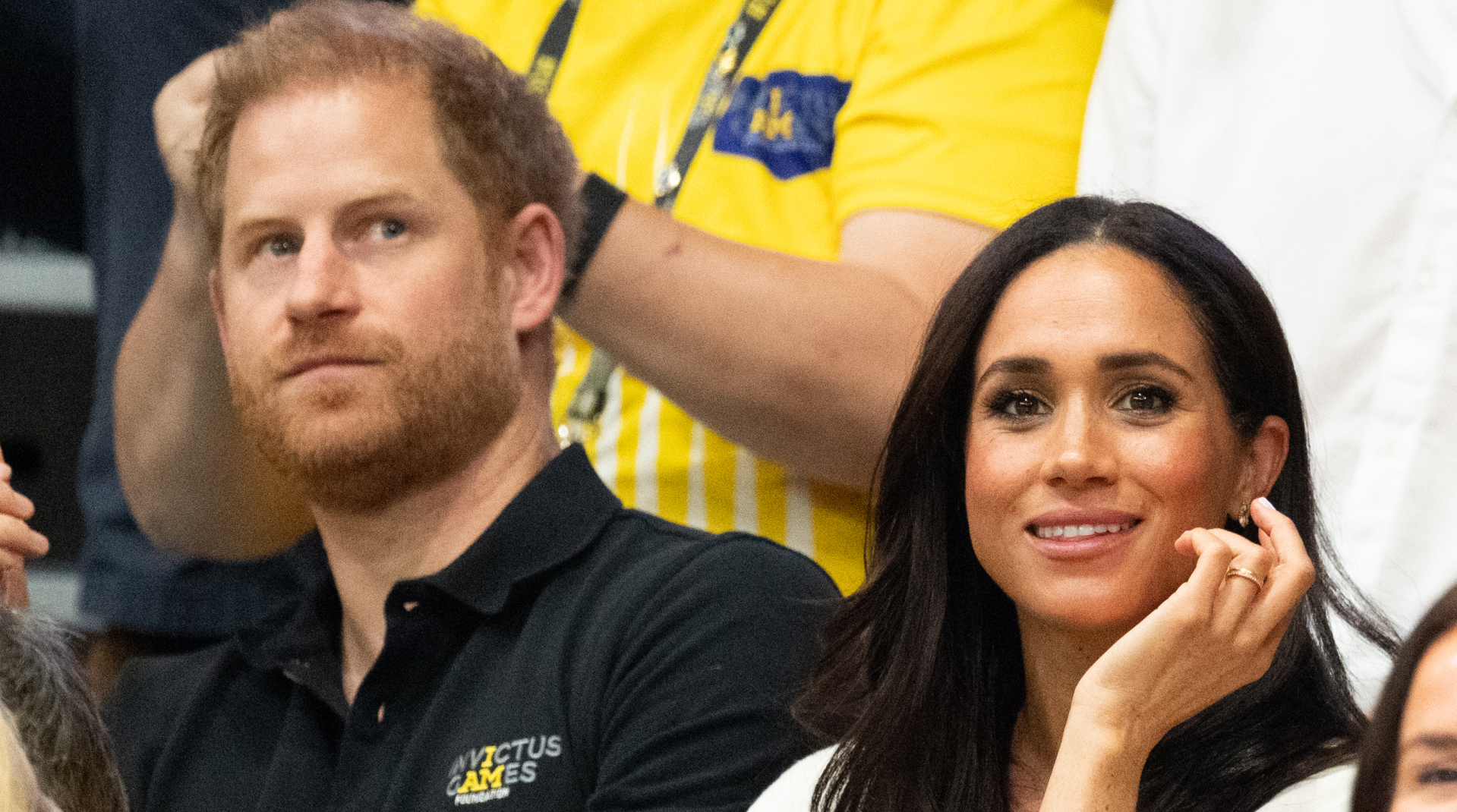 Meghan Markle wears a white jacket and smiles and has long black hair