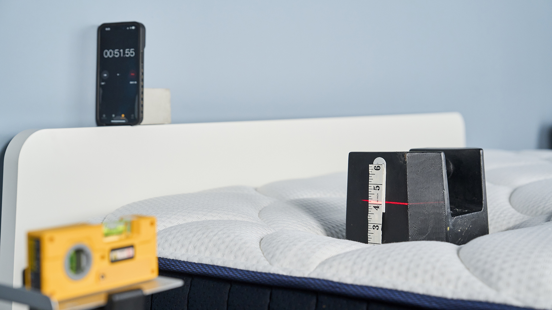 A 25kg weight on the DreamCloud mattress with a measuring tape along its side. A laser red line points at the weight, showing the weight has sank over 4". In the background a phone shows a stopwatch.