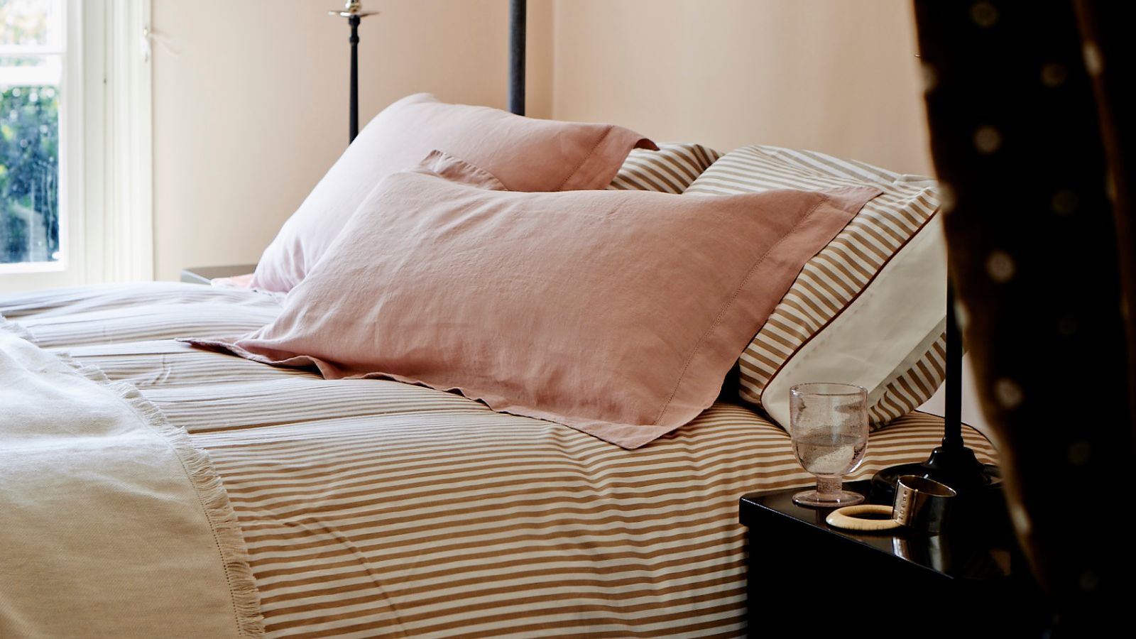 A peach bedroom with striped pink bedding, pink pillows, and a dark brown and white polka dot curtain in the foreground.