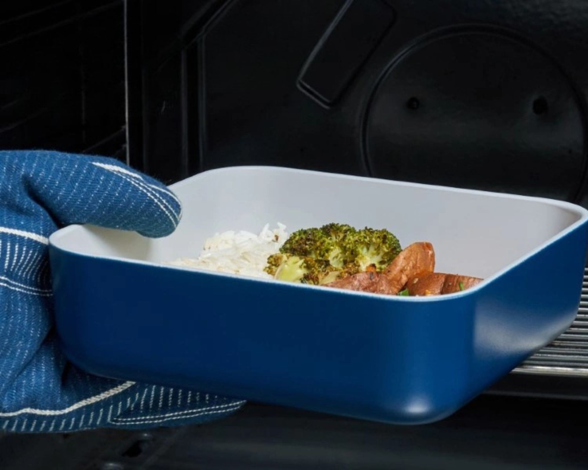 Non-stick Caraway food container in dark blue color being taken out of the oven with oven gloves. The container has rice, broccoli, and chicken