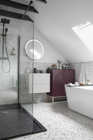 Bathroom with terrazzo wall and floor tiles, walk-in black framed shower, white bath, grey wall-hung vanity, round mirror and purple cabinet