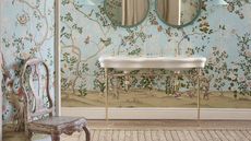 Bright, large bathroom with pale blue scene wallpaper feature and Victorian double sink with gold hardware, and a vintage, distressed chair