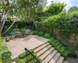 An example of how to lay a patio with steps leading up to a shaded patio area with chairs, small table and deep flower beds.