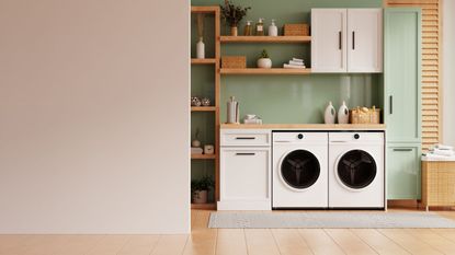 A sage and pink laundry room with wooden shelving and shutters, two washing machines, white cabinets, diffusers, plants and bottles and a grey rug on a brown wooden floor, with a washing basket to the right