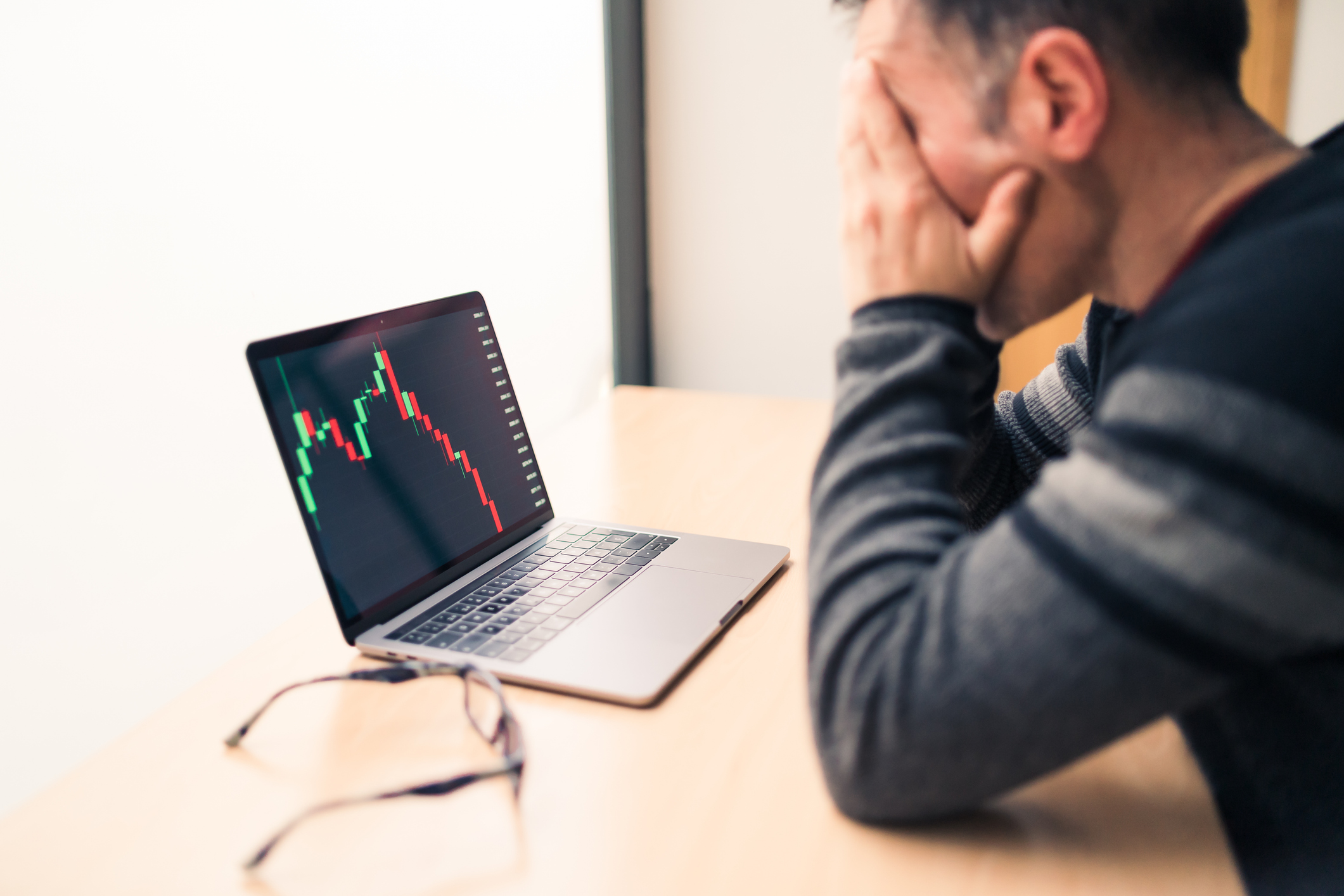 An older man covers his eyes in distress as his laptop shows a graph going down, ostensibly a falling 401(k) in a down market.