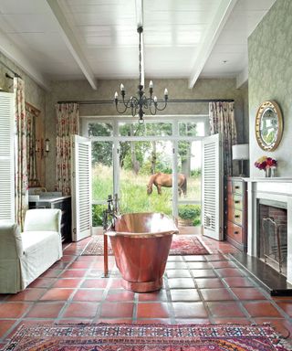 bathroom with copper bath, white chaise, shutters, green patterned wallpaper, a chandelier and terracotta floor tiles