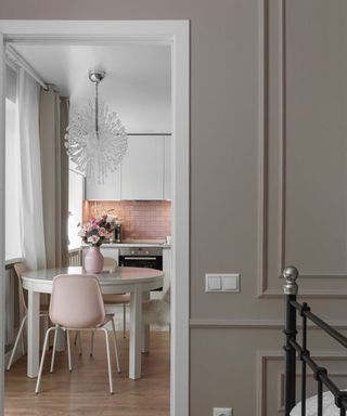 A dining idea for a small kitchen with glass chandelier, pink metro tile backsplash, white round table, pink vase with flowers, pink chairs and taupe curtain window treatment