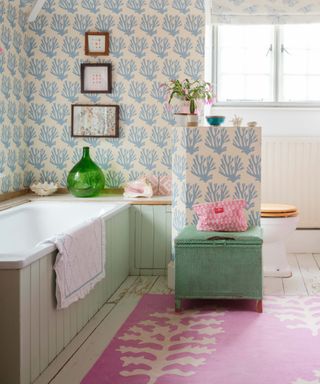 Pastel colored bathroom with blue coral wallpaper, pastel green bath panels, pink rug, shells, plants, artwork