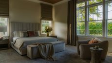 Image of a neutral and gray, modern bedroom design. There is a large gray bed in the middle with minimal bedding. There are two windows on either side of the bed with curtains and a large window to the right with curtains as well.