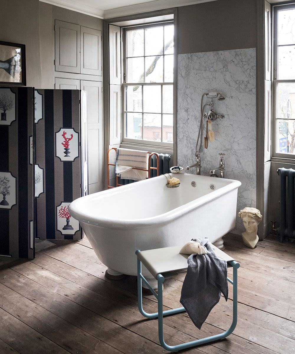 A bathroom with wooden flooring, a white free-standing bath and two sash windows